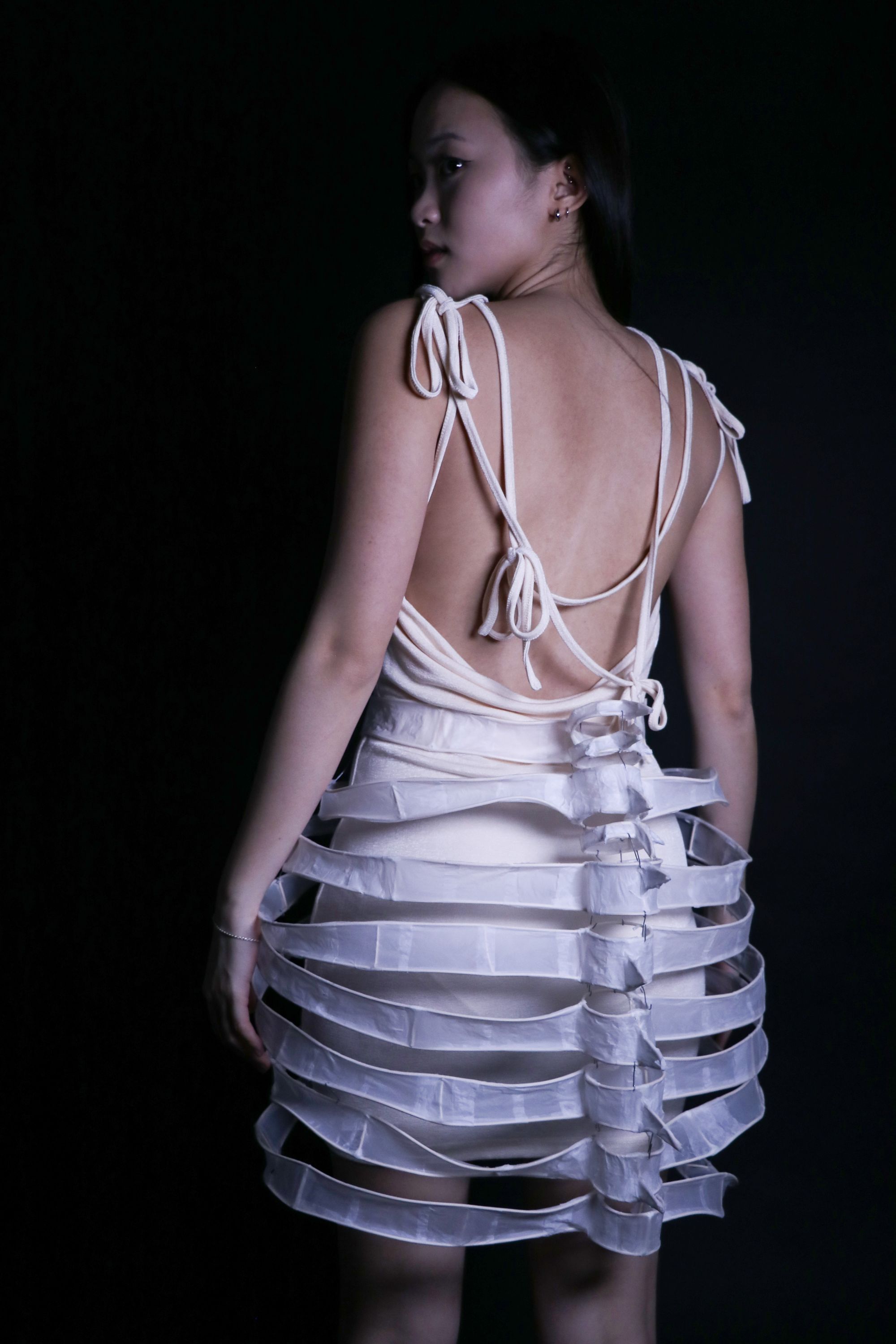 Model stands in profile against a dark background wearing a white dress with an open back and strappy shoulder ties. The dress features a dramatic cage-like skirt structure made of translucent bands resembling ribs.